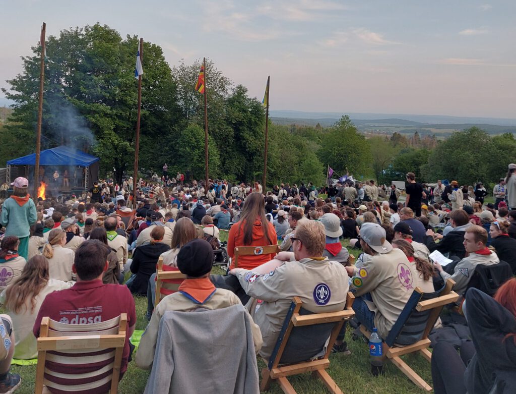 Überblick über den Rand der Arena. Viele Menschen sitzen auf Schwedenstühlen und schauen Richtung Bühne.