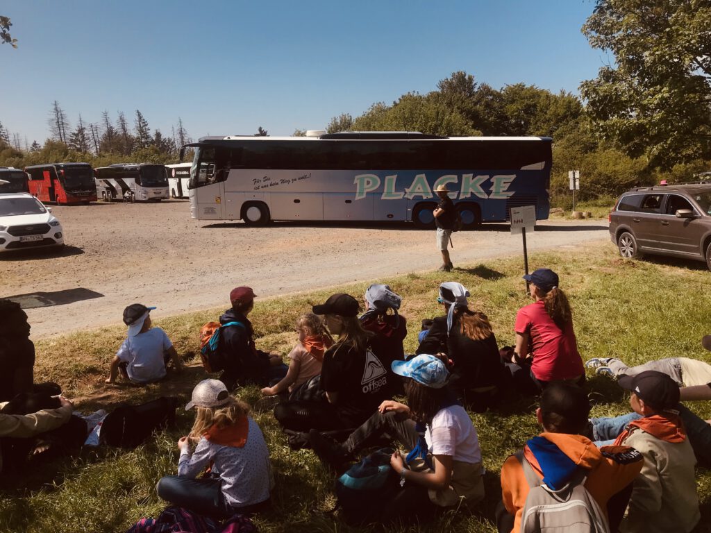 Die Kinder sitzen im Schatten und warten auf den Bus. Dahinter ist der Reisebus der Firma Placke auf dem Parkplatz zu sehen.