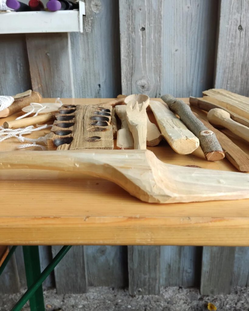 Einige aus Holz geschnitzte Gegenstände liegen auf einem Tisch: ein Tortenheber, mehrere Löffel und ein Feuerbohrer